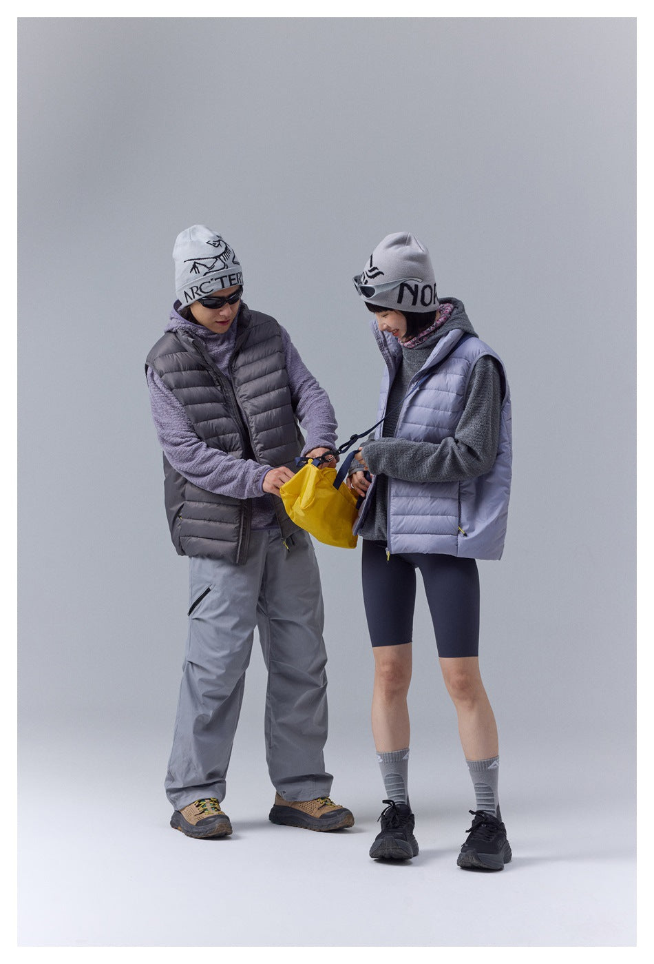 Two models in charcoal and pale lavender quilted vests look down at a yellow shoulder bag.