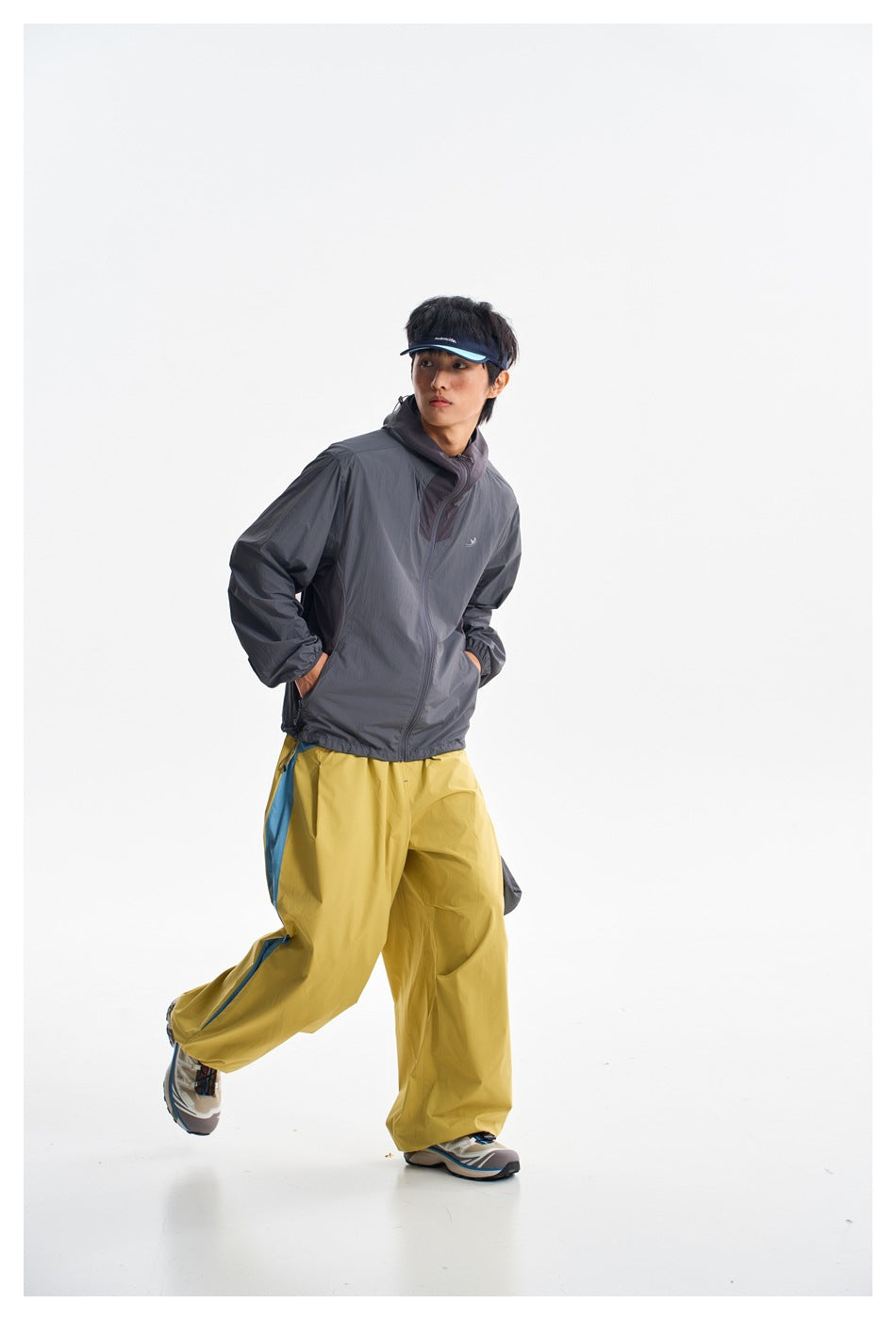 A person poses in a charcoal hooded jacket and mustard-yellow pants with blue side panels, one leg lifted.