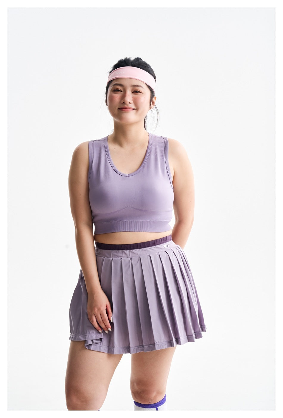 Closer front view of a model wearing a lavender V-neck sports bra and pleated skirt with a dark waistband.