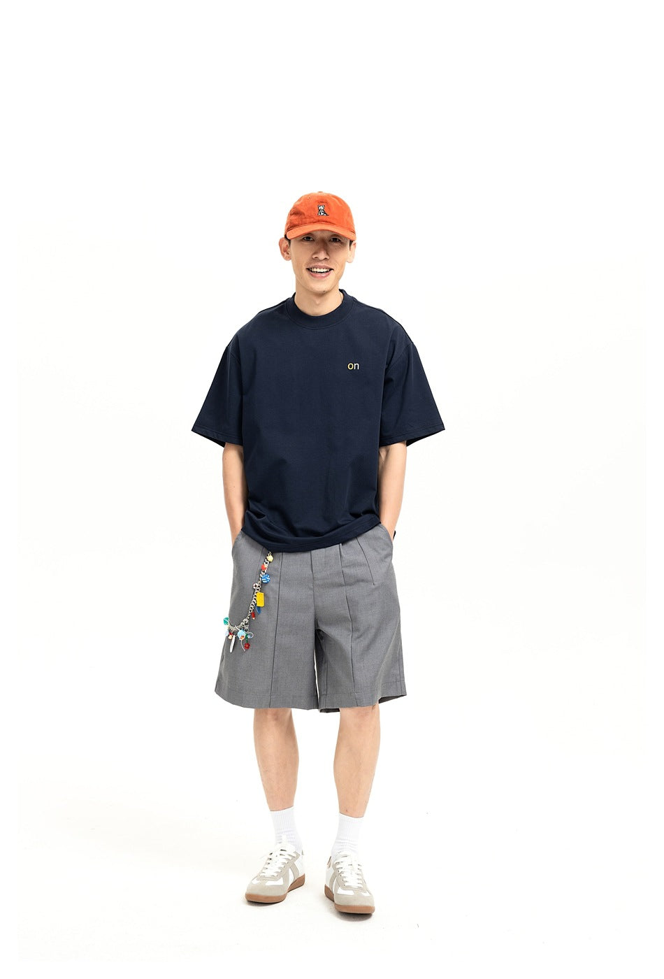 Model wearing a navy oversized T-shirt with a small chest mark, gray pleated shorts, and an orange cap.