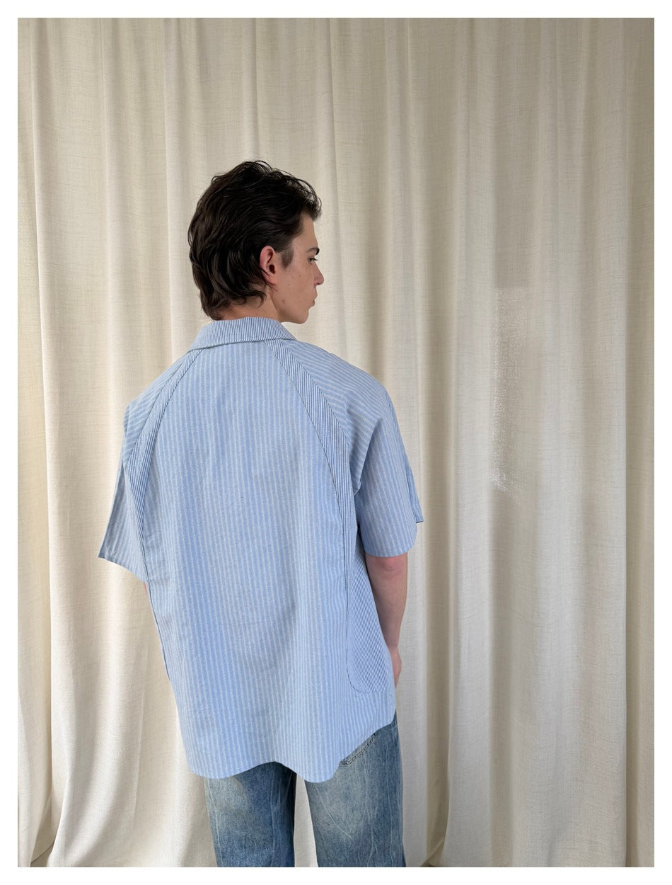 Back view of a person wearing a loose light blue striped short-sleeve shirt with contrast panels and curved side pockets.