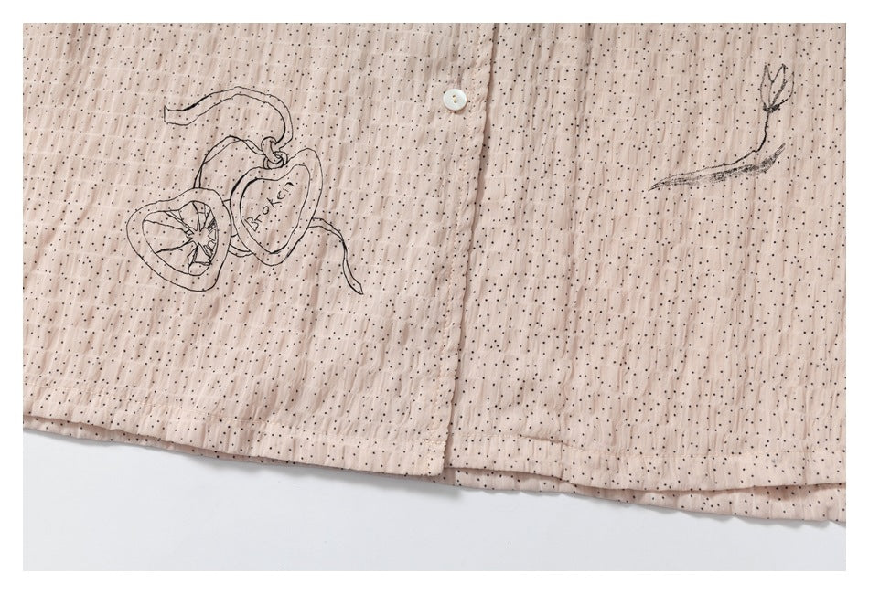 Lower garment detail showing a button, dotted blush fabric, and fruit and feather illustrations.