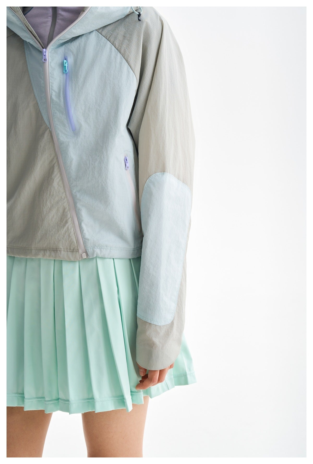 Close-up of a gray and pale blue jacket with contrast zippers and elbow panels, worn over a mint pleated skirt.