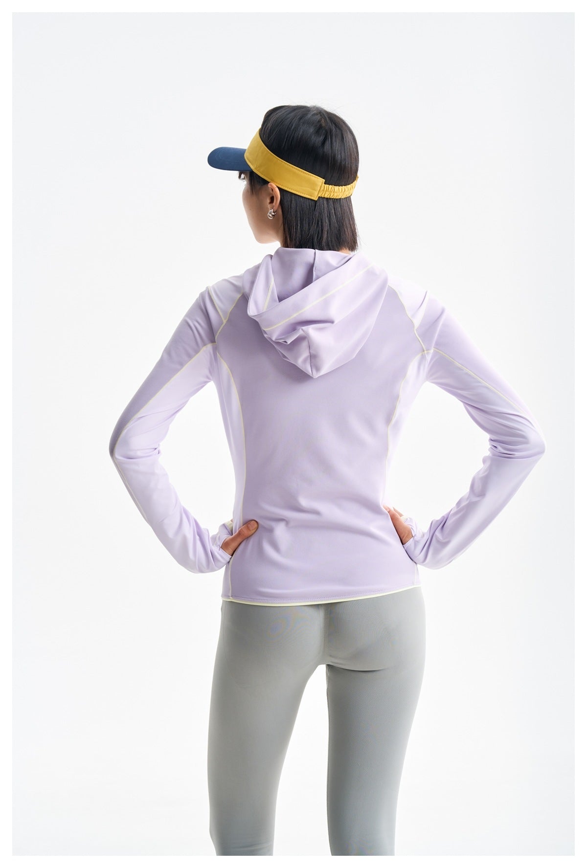 Rear view of a model wearing a lavender hooded top with pale yellow seam accents and gray leggings.