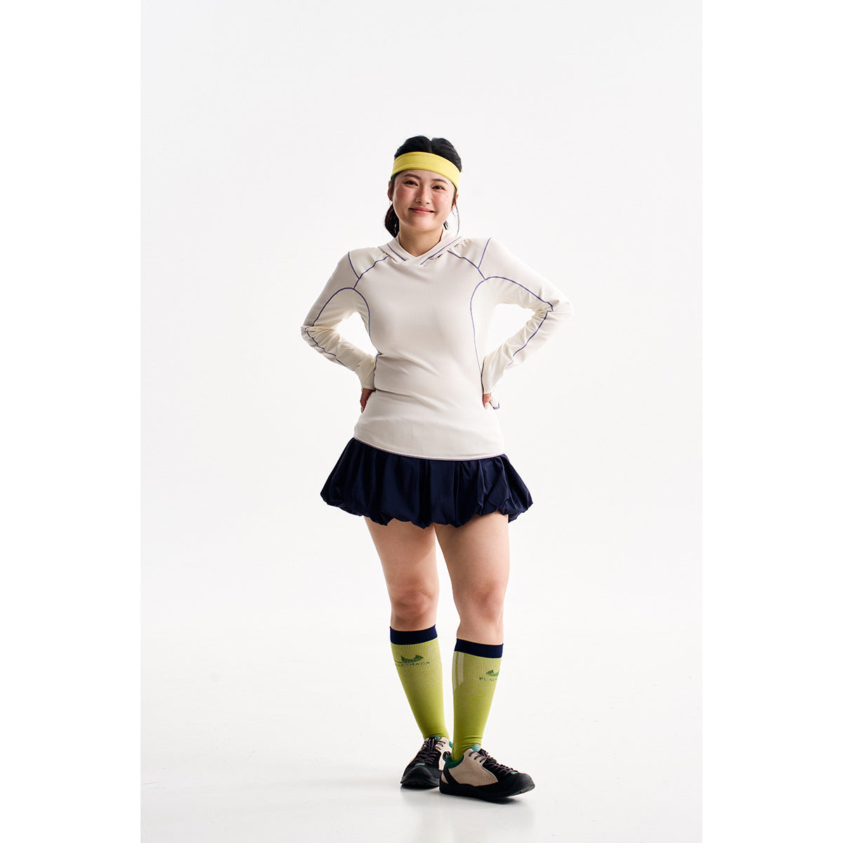 Model wearing a white hooded athletic top, navy bubble skirt, yellow-green knee socks, and a yellow headband.