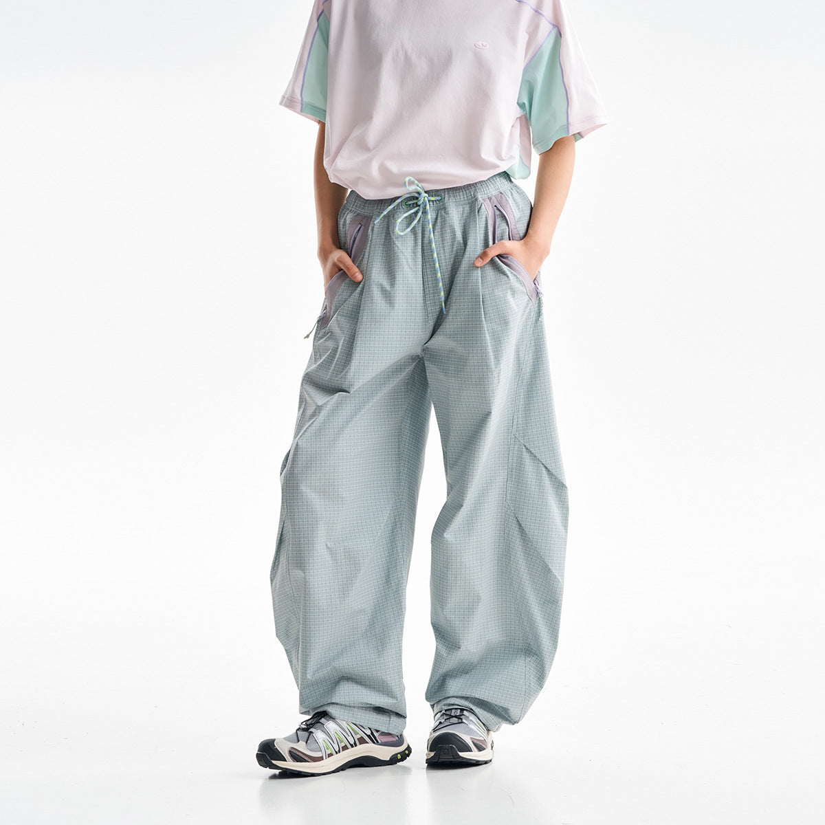 Front view of loose pale gray-green plaid pants with a drawstring waist and angled pockets, worn with a color-block shirt.