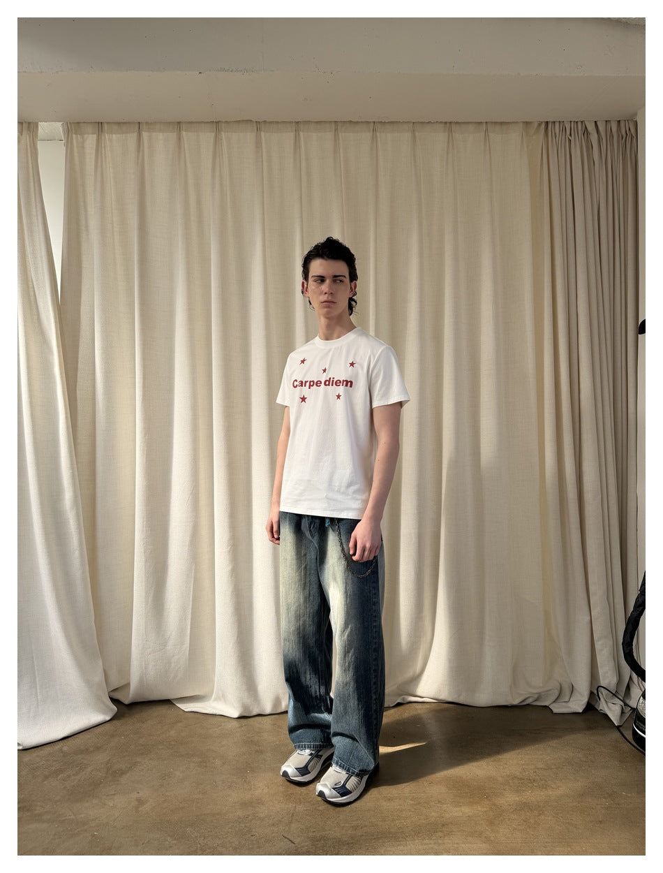 Person wearing a white T-shirt with red star and 'Carpe diem' print, faded wide-leg jeans and sneakers in front of cream