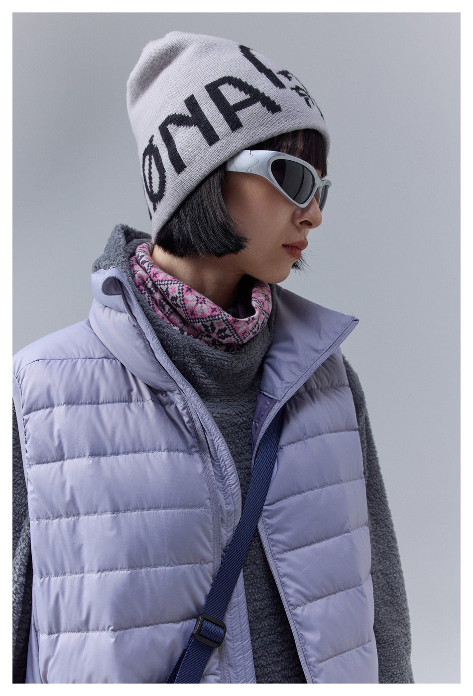 Close view of a model in a pale lavender quilted vest, charcoal fleece, patterned neck warmer, and white graphic beanie.