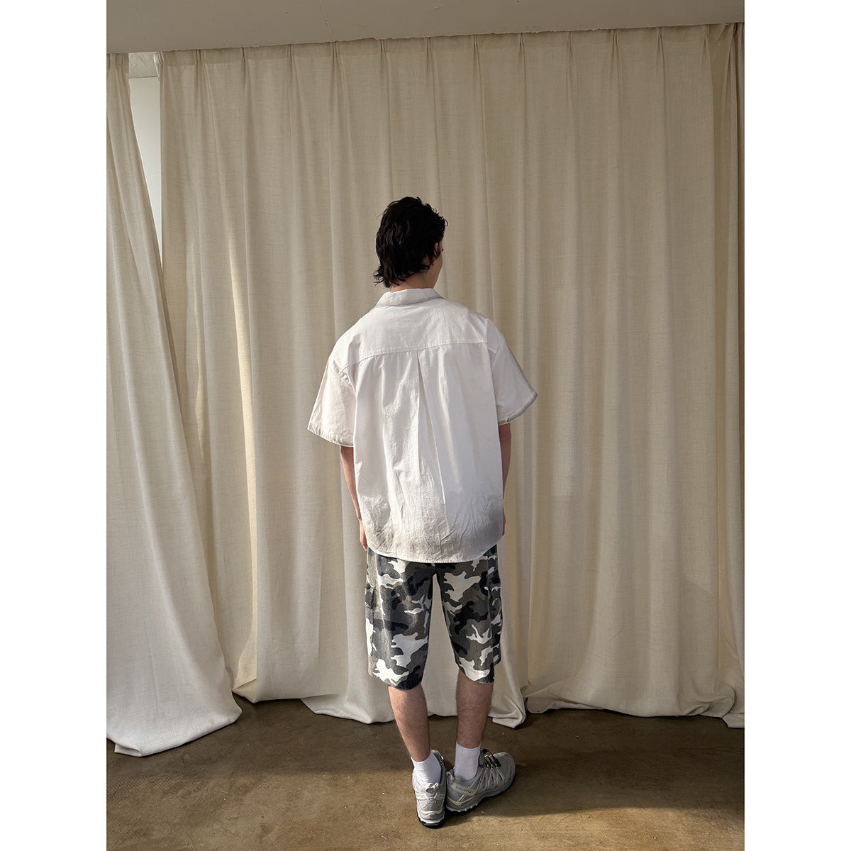 Back view of a model wearing a white short-sleeve shirt and gray camouflage cargo shorts.