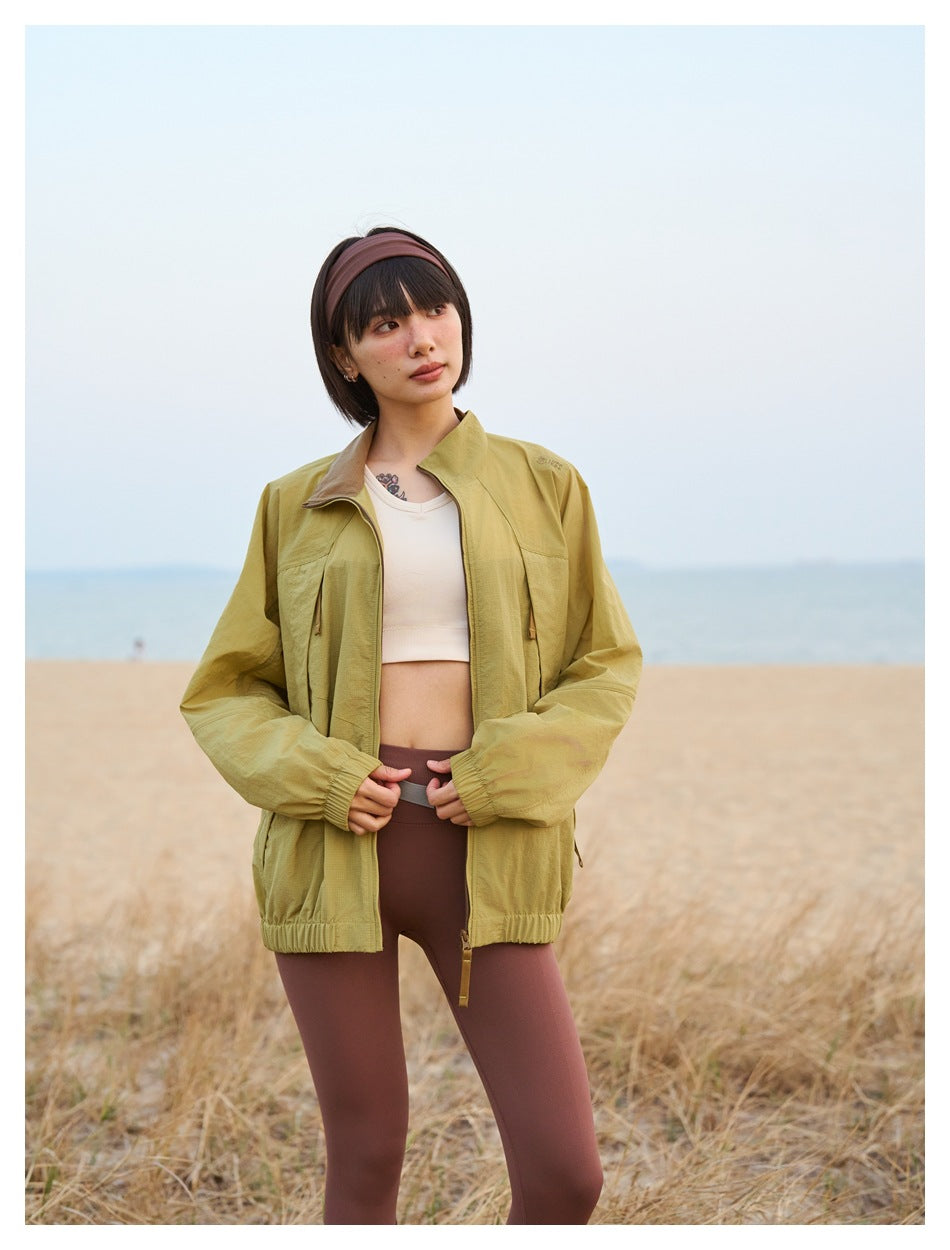 Model wearing an open mustard-olive zip jacket with elastic cuffs and hem over a pale cropped top.