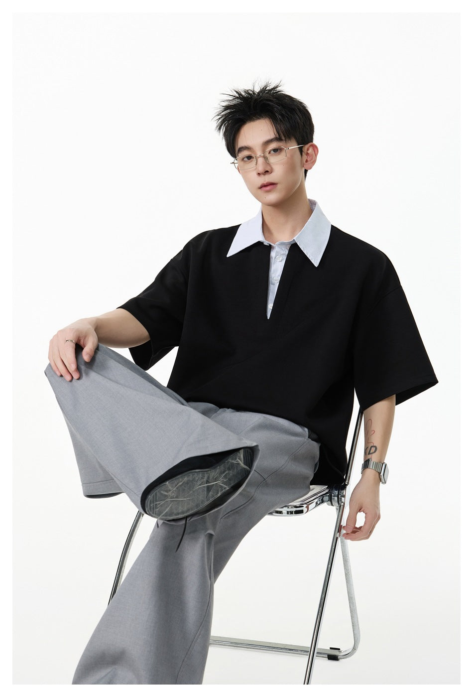 Seated model wearing a black oversized short-sleeve shirt with a white pointed collar and gray trousers.