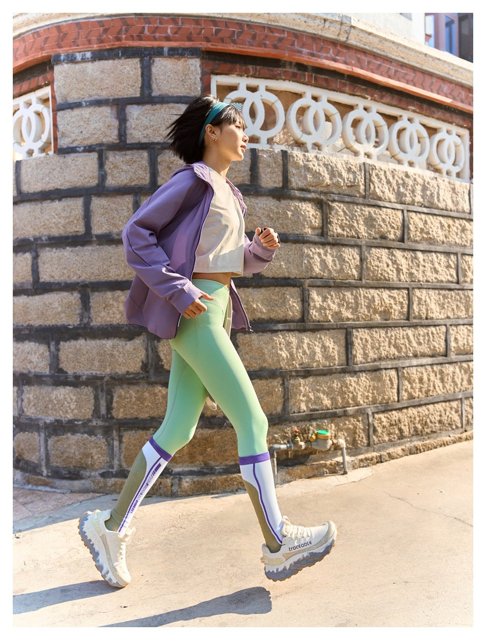 Woman walking in a lilac jacket, white crop top, mint leggings, and tall white-and-purple socks.