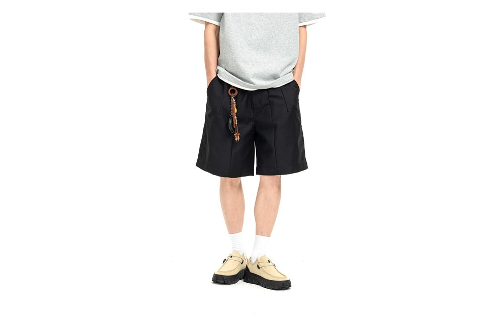 Cropped model wearing black pleated shorts with a dangling accessory, a gray T-shirt, and beige shoes.