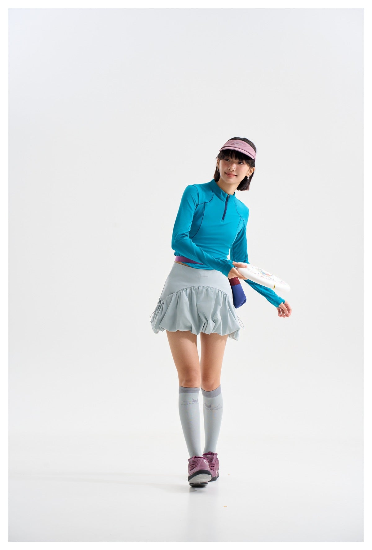Model wearing a teal quarter-zip top and pale blue-gray gathered skirt, holding a white paddle.