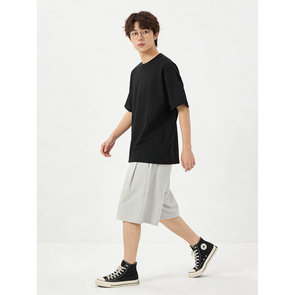 Model wearing an oversized black T-shirt with a small chest emblem and light gray shorts, shown from the front.