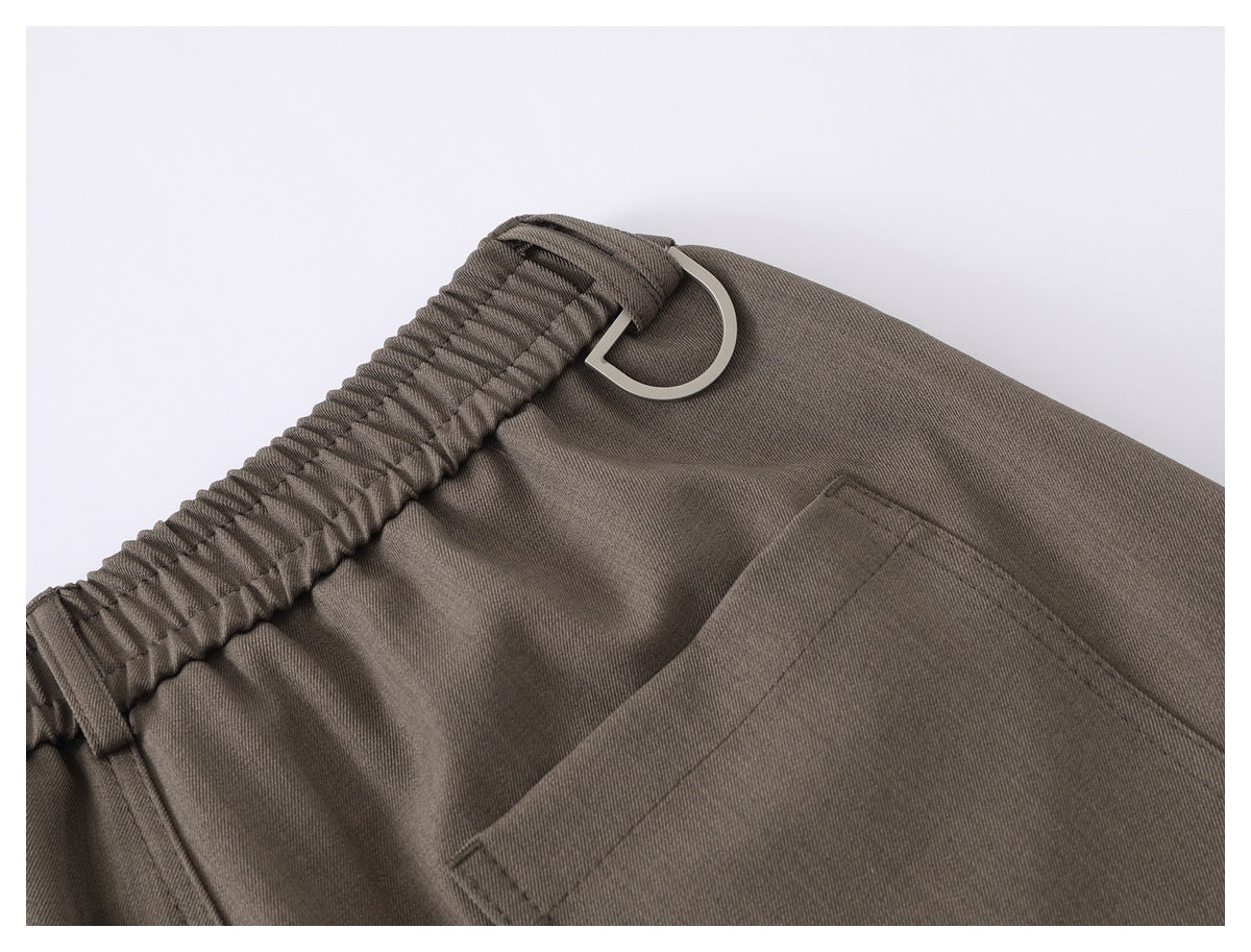 Close-up of taupe-gray pants showing an elastic waistband, silver D-ring, and stitched pocket.