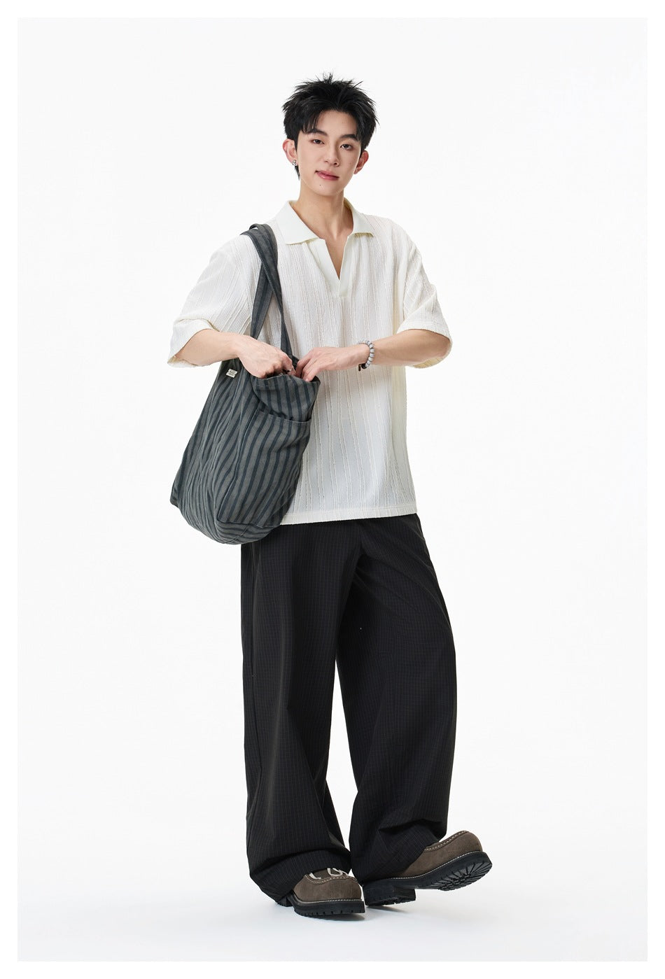 Model wearing an ivory short-sleeve polo with vertical texture and wide black trousers, carrying a dark striped tote.