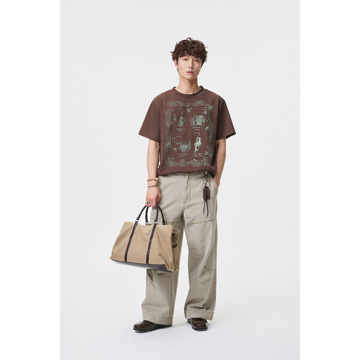 Full-length model in a brown graphic T-shirt and loose beige trousers, carrying a tan tote bag.