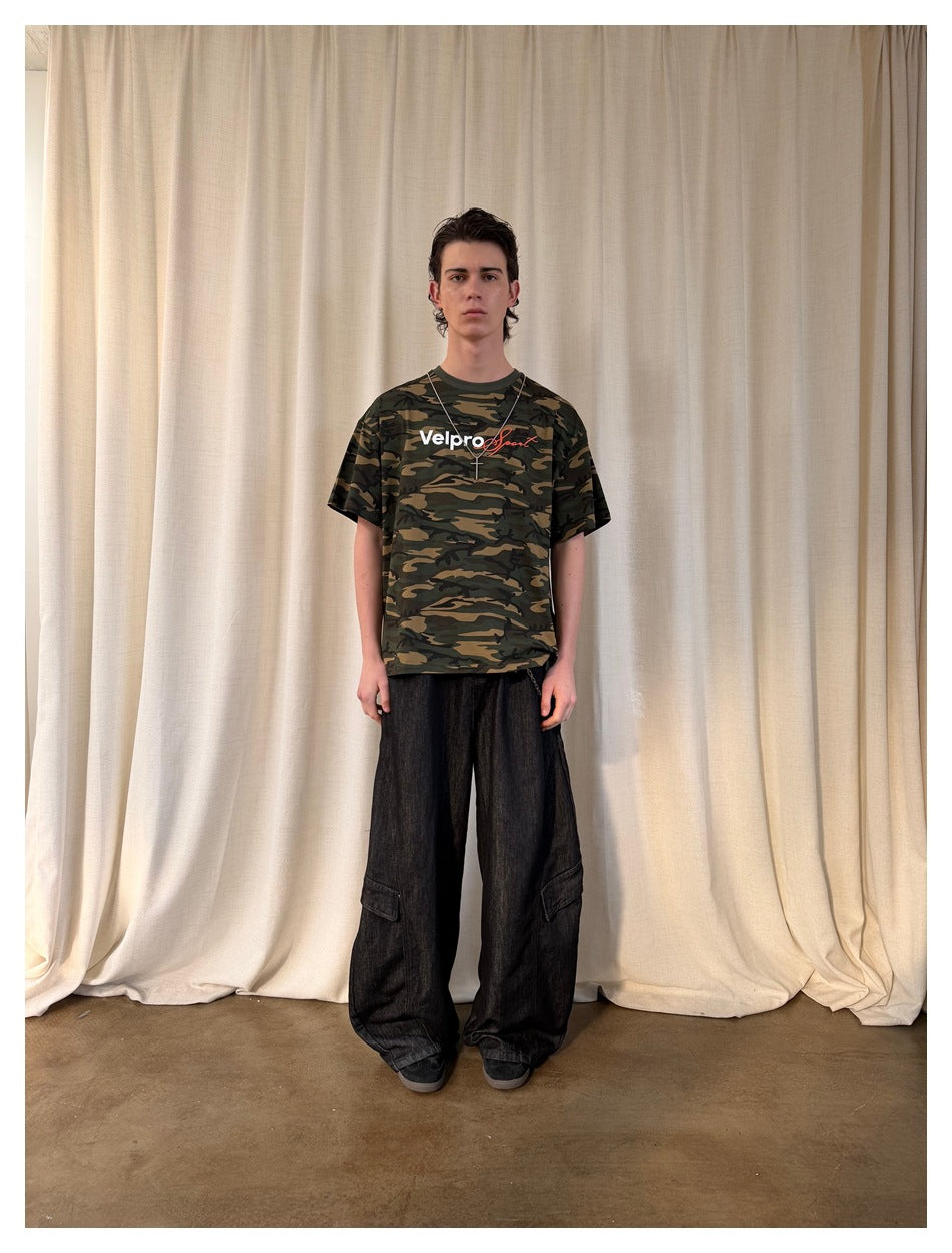 Full-length front view of a model in a green and brown camouflage graphic T-shirt and loose black cargo jeans.