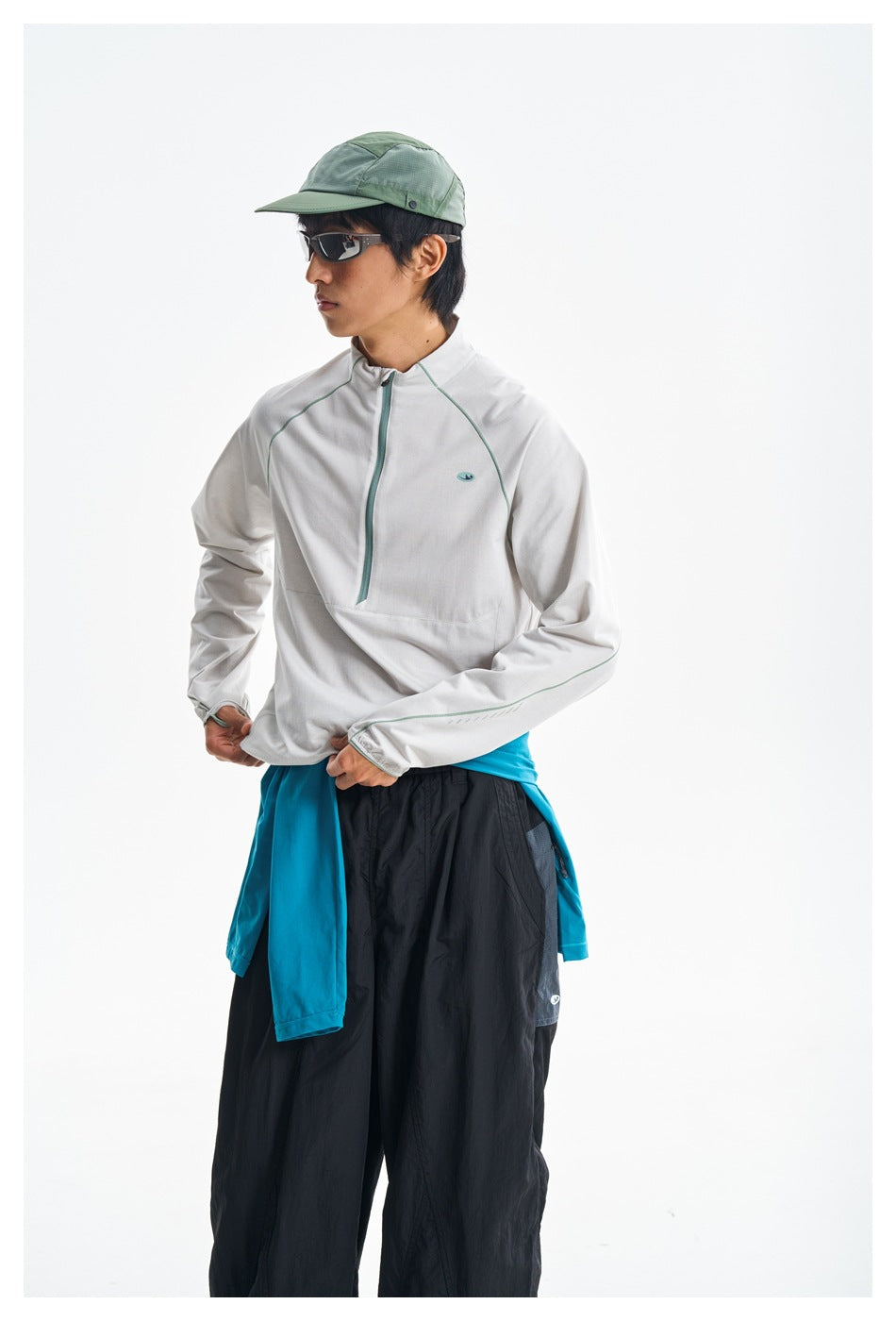 Model wearing a white pullover with a teal quarter zip and piping, black pants, and a turquoise layer tied at the waist.