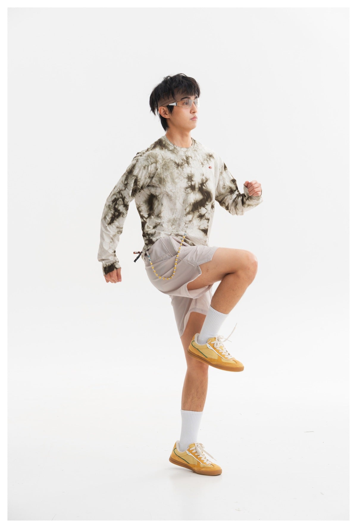 Model in glasses wearing an olive-gray tie-dye long-sleeve shirt and pale gray shorts, posed with one knee raised.