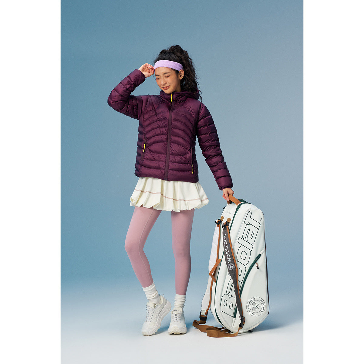 A woman models a plum hooded puffer jacket with a white skirt and pink leggings beside a tennis bag.