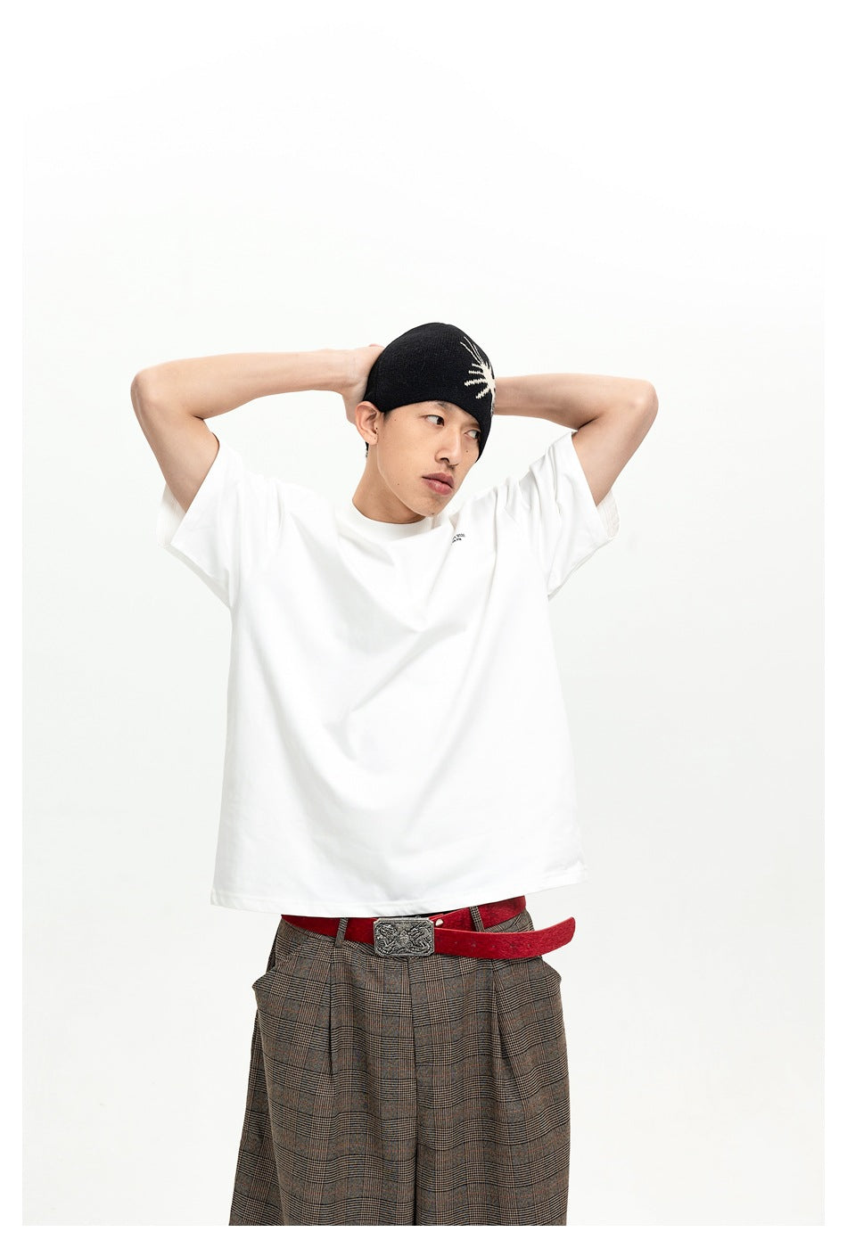Model wearing a white oversized T-shirt and brown plaid trousers, shown from the waist up with hands behind his head.