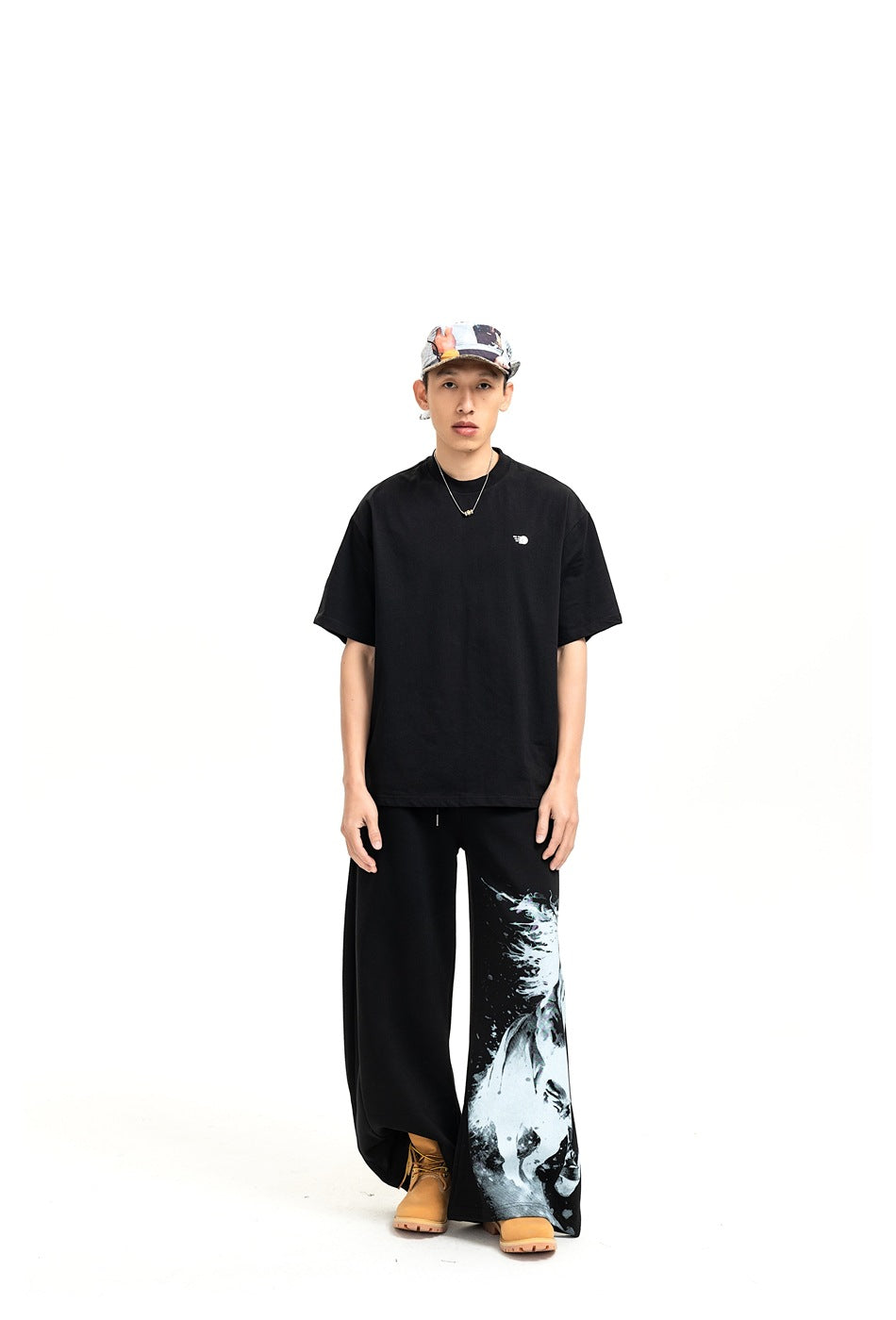 Model wearing black wide-leg sweatpants with a horse graphic on the lower leg, a black tee, cap and tan boots