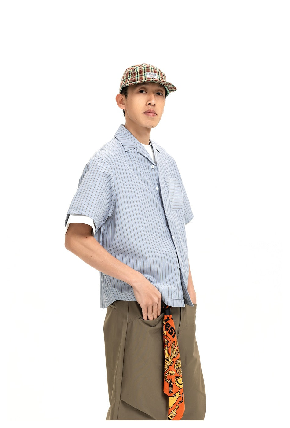 Model wearing a light blue pinstriped short-sleeve shirt with a chest pocket, olive trousers, and a plaid cap.