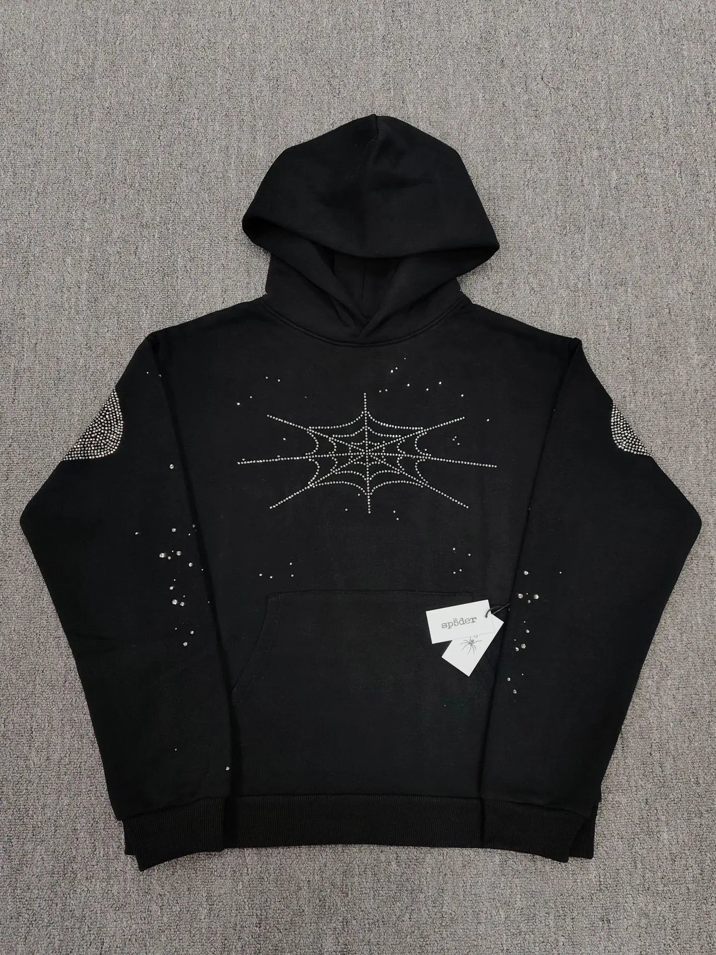 Sp5der black pullover hoodie with silver rhinestone spider web design on chest and shoulders laid flat on grey surface