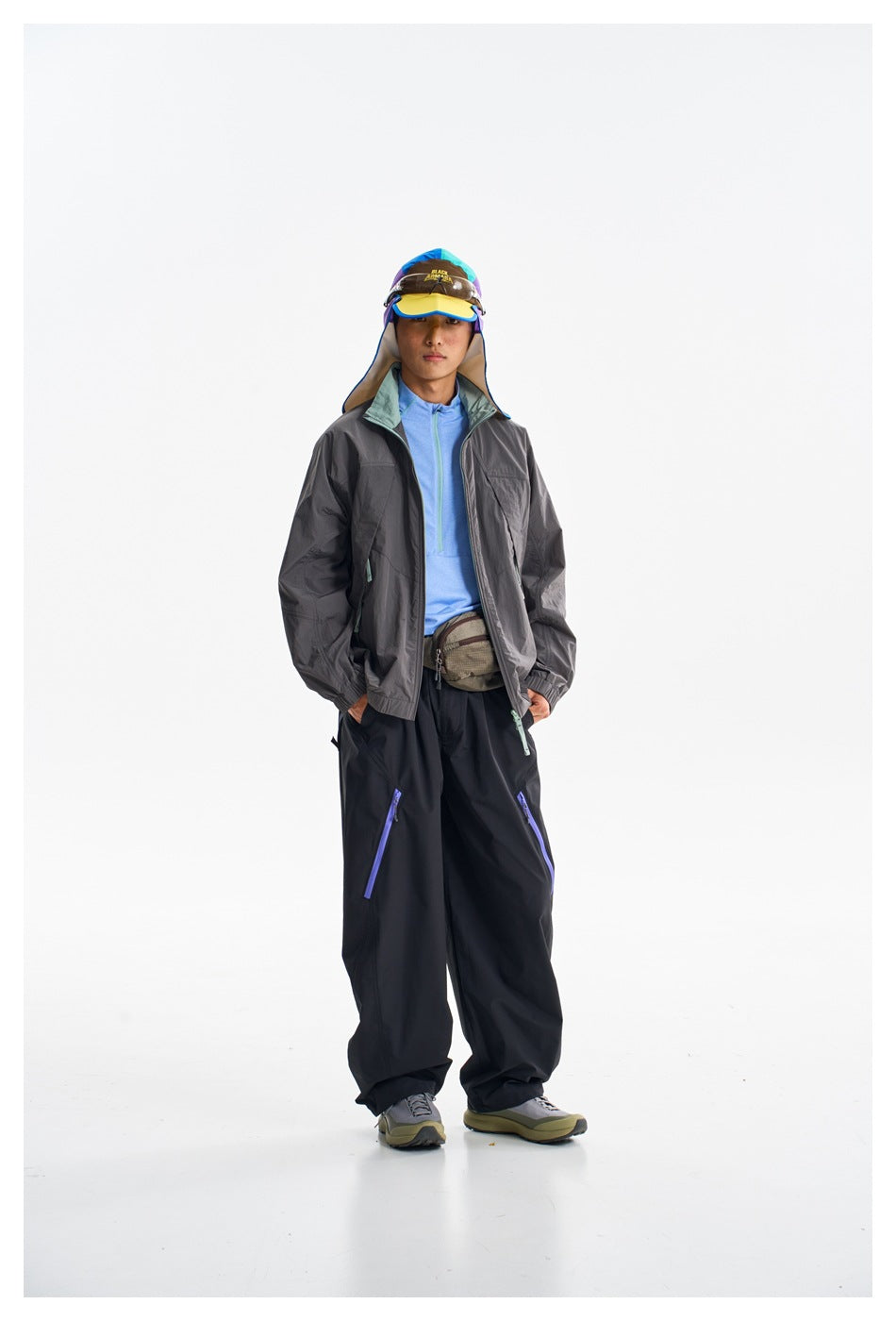 Full-length model wearing a charcoal jacket, blue top, black trousers, waist pack and sun hat.