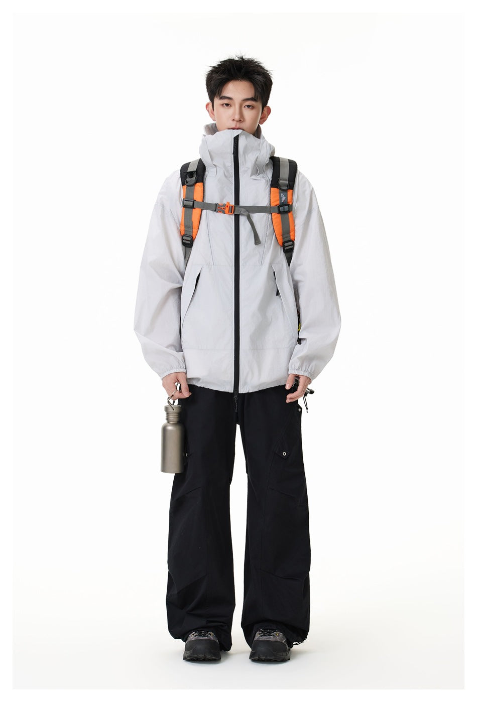 Full-length front view of a model wearing a light gray hooded jacket, orange-and-gray backpack, and loose black pants.