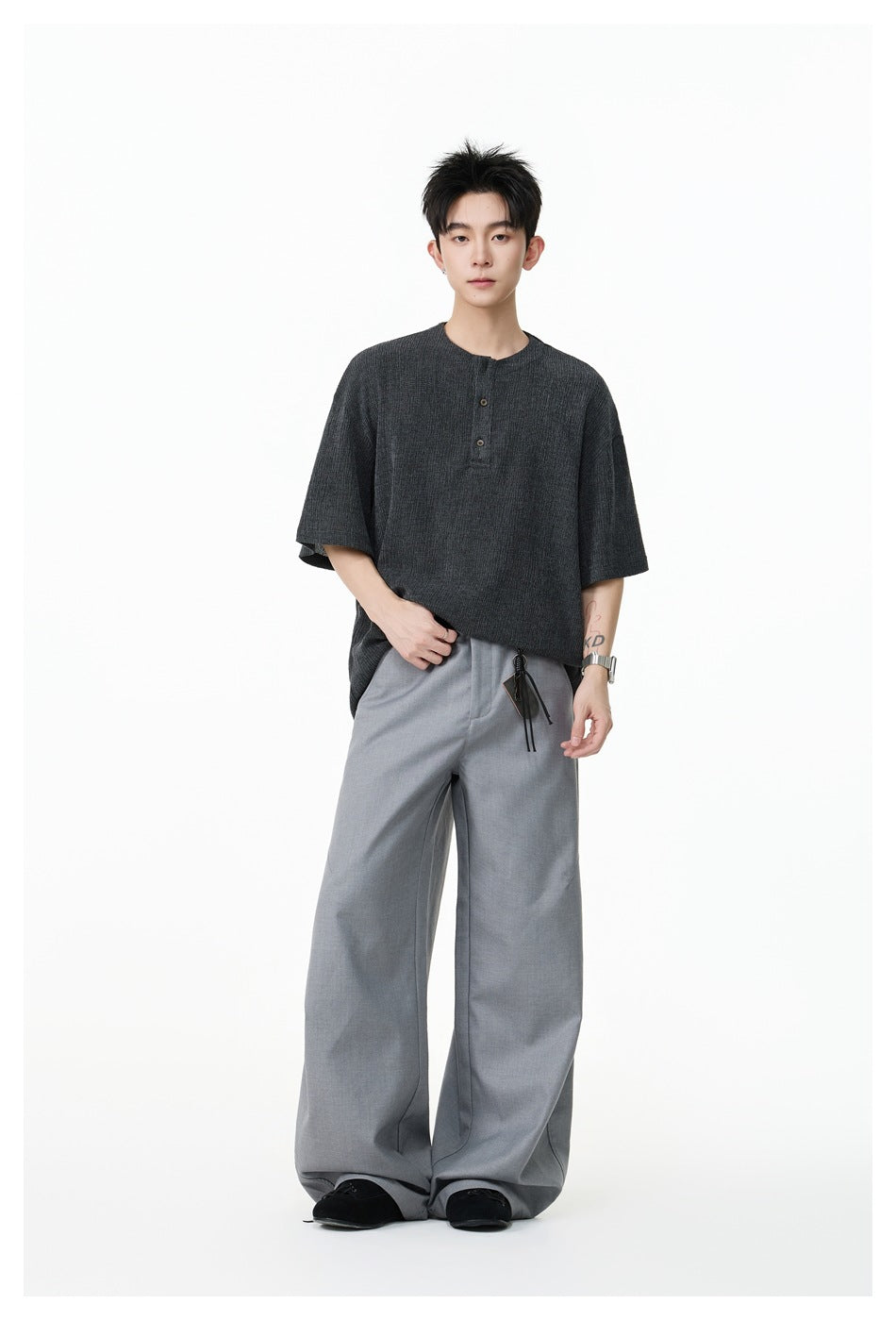Model wearing a charcoal oversized short-sleeve button-placket top and gray wide-leg trousers, with one hand at the hem.