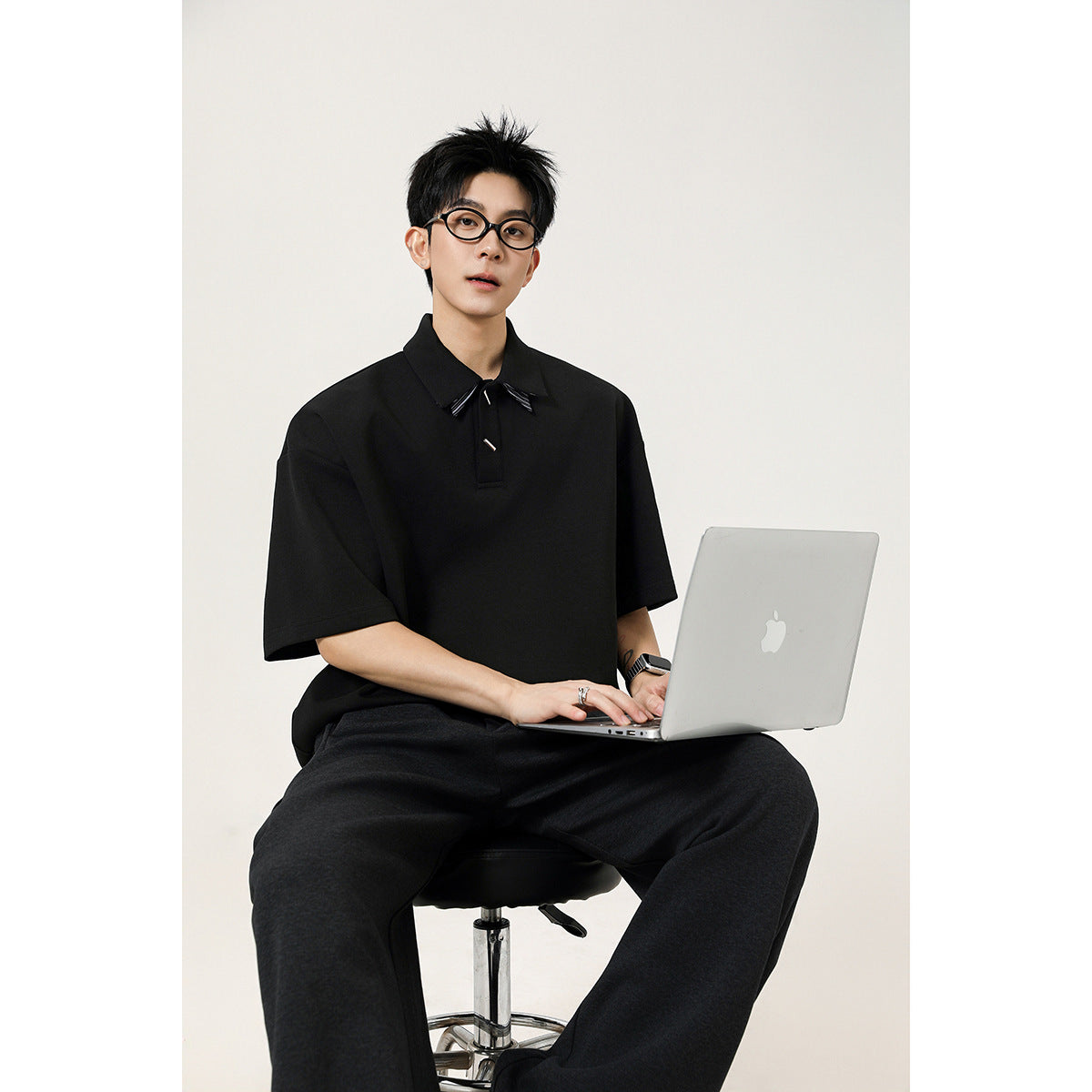 Model seated in a black oversized short-sleeve polo with a pointed collar and small decorative accents.