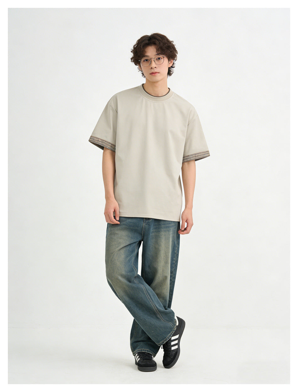 Model wearing a light beige short-sleeve crew-neck T-shirt with patterned sleeve cuffs and loose blue jeans.