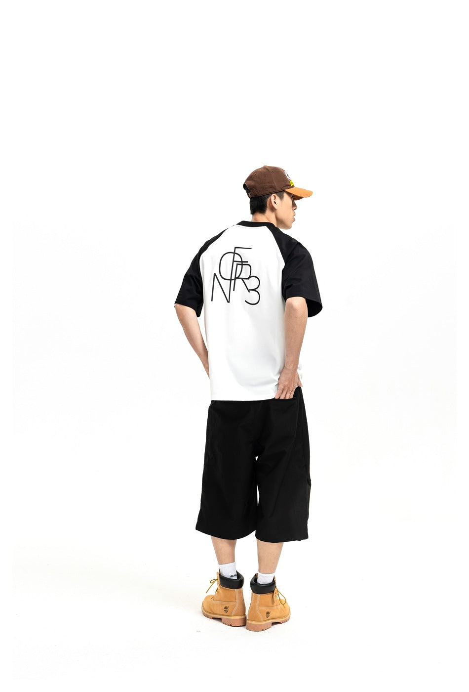Model seen from behind wearing a white raglan T-shirt with black sleeves and a black back graphic, with black shorts and a