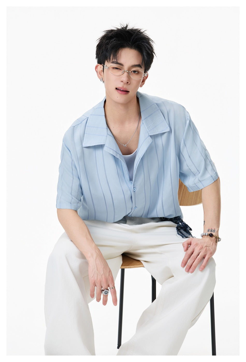 Seated model wearing a light-blue vertical-stripe short-sleeve shirt and white trousers.