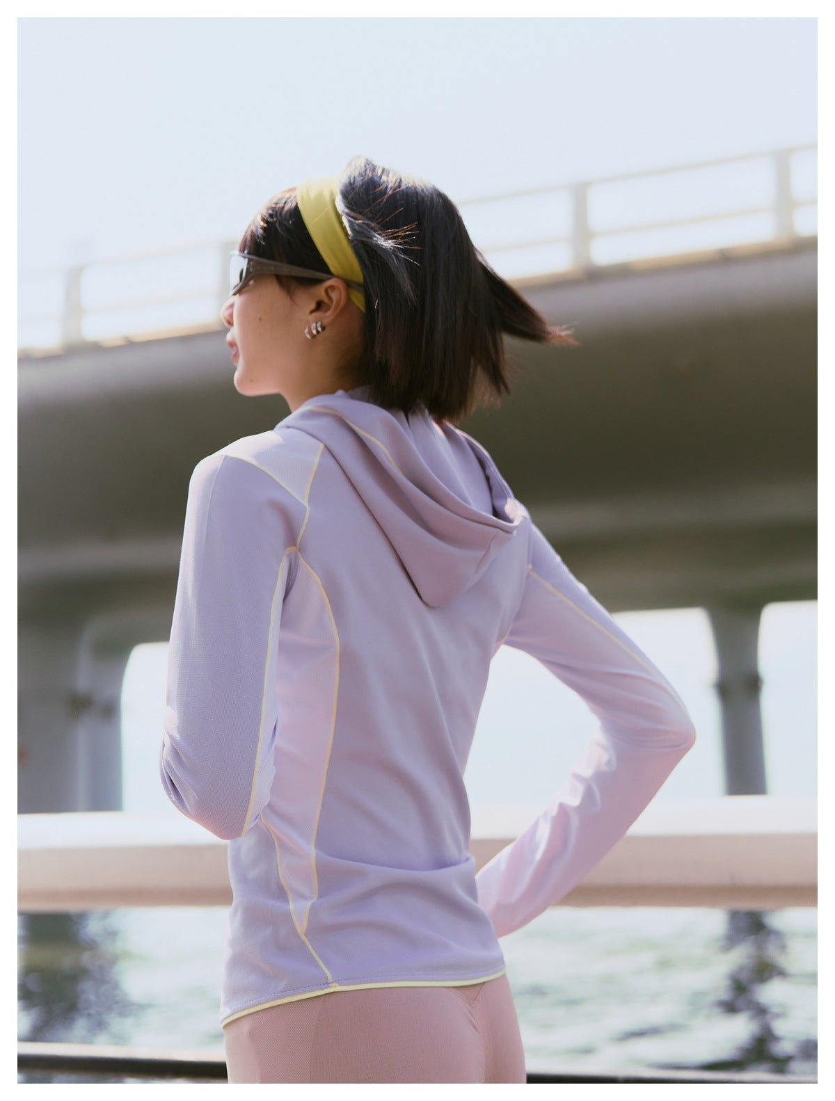 Rear view of a model wearing a lavender hooded athletic top with pale yellow piping outdoors.