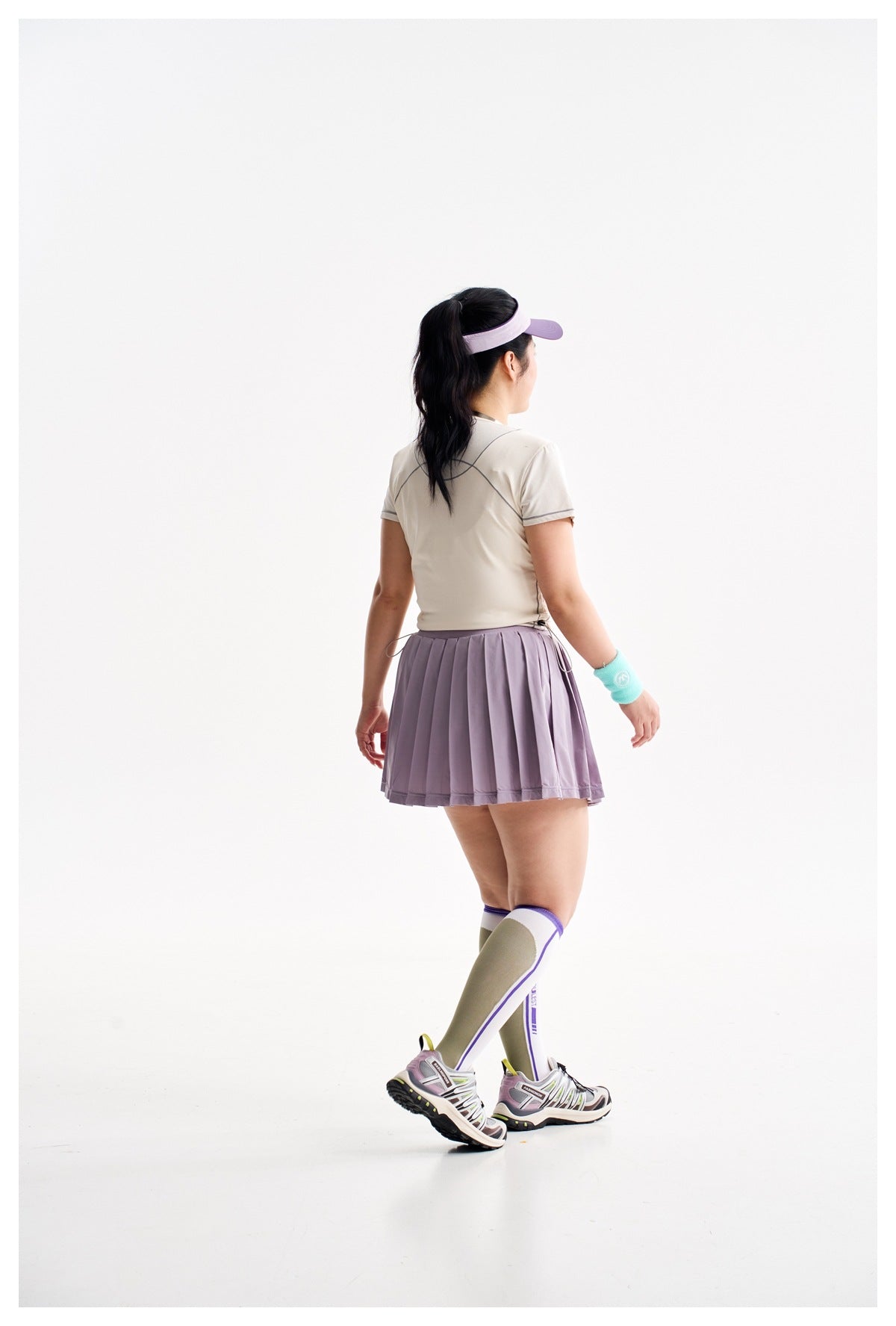 Back view of a model wearing a pleated lavender athletic skirt, light top, knee-high socks, and sneakers.