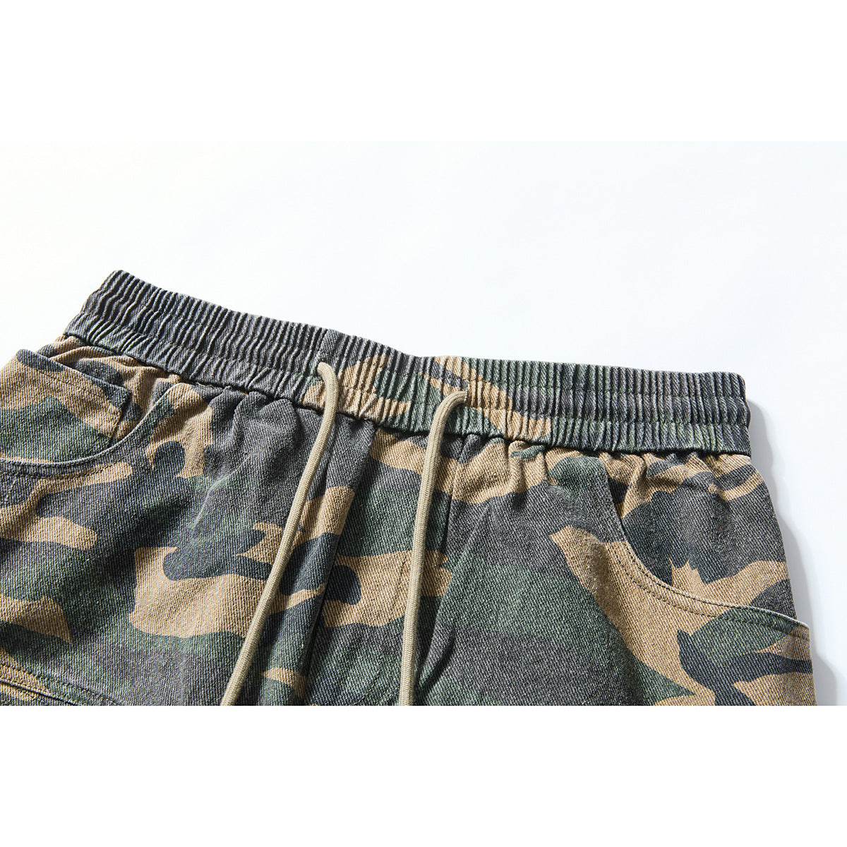 Close-up of camouflage shorts with an elastic waistband, tan drawstrings, and a curved front pocket.