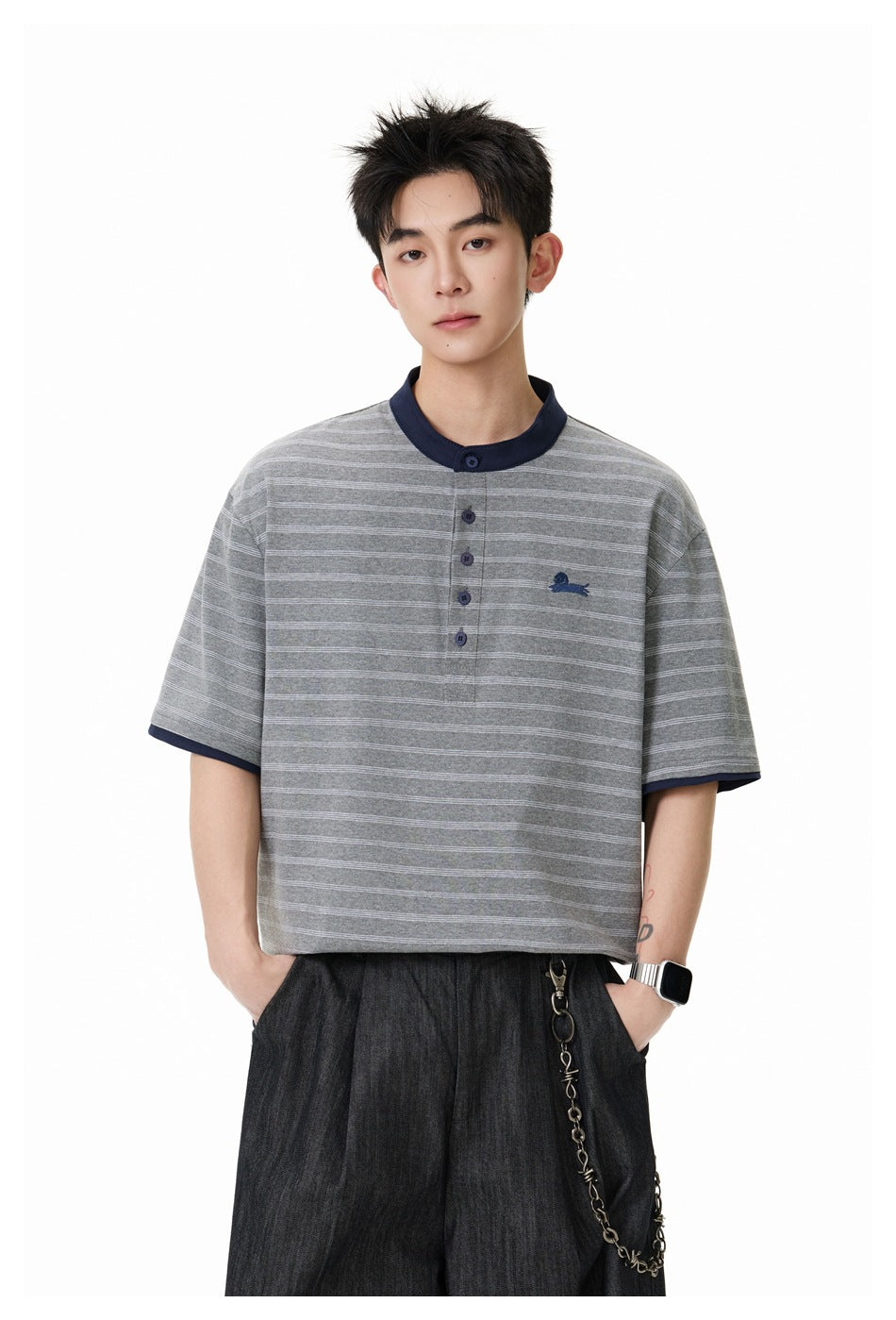 Model wearing a gray striped short-sleeve henley with navy trim and dark trousers, hands in pockets.