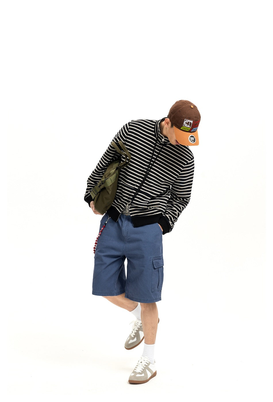 Model leaning forward in blue cargo shorts, striped jacket, cap and olive bag against a white background