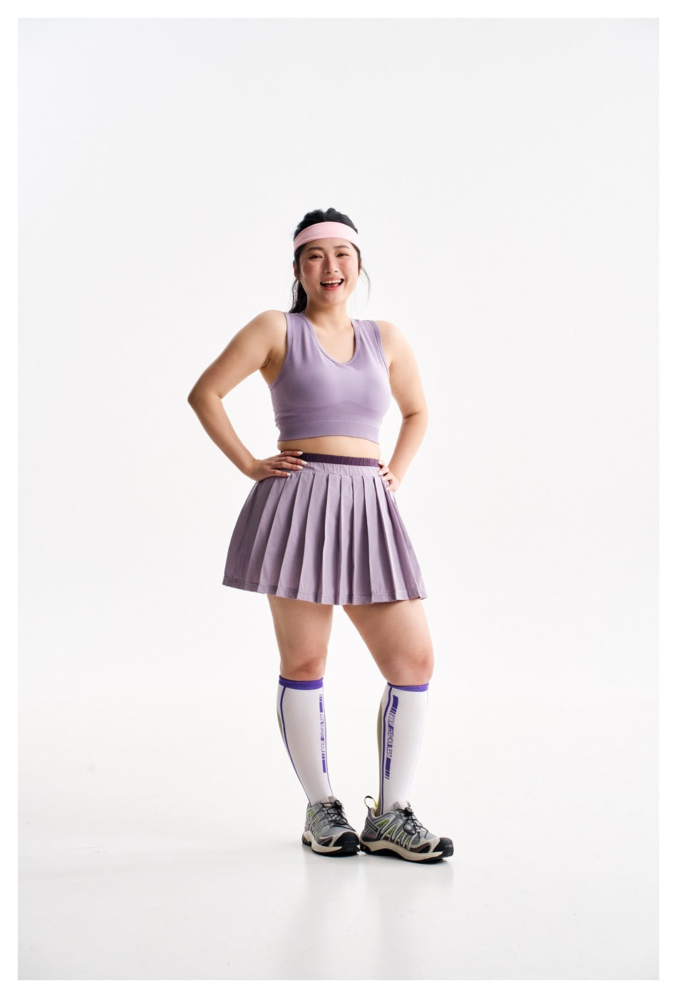 Front view of a smiling model in a lavender sports bra and pleated skirt, posing with hands on her hips.