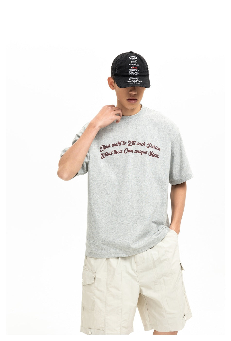 Model in a light gray graphic T-shirt and cream cargo shorts, touching the collar while wearing a black cap.