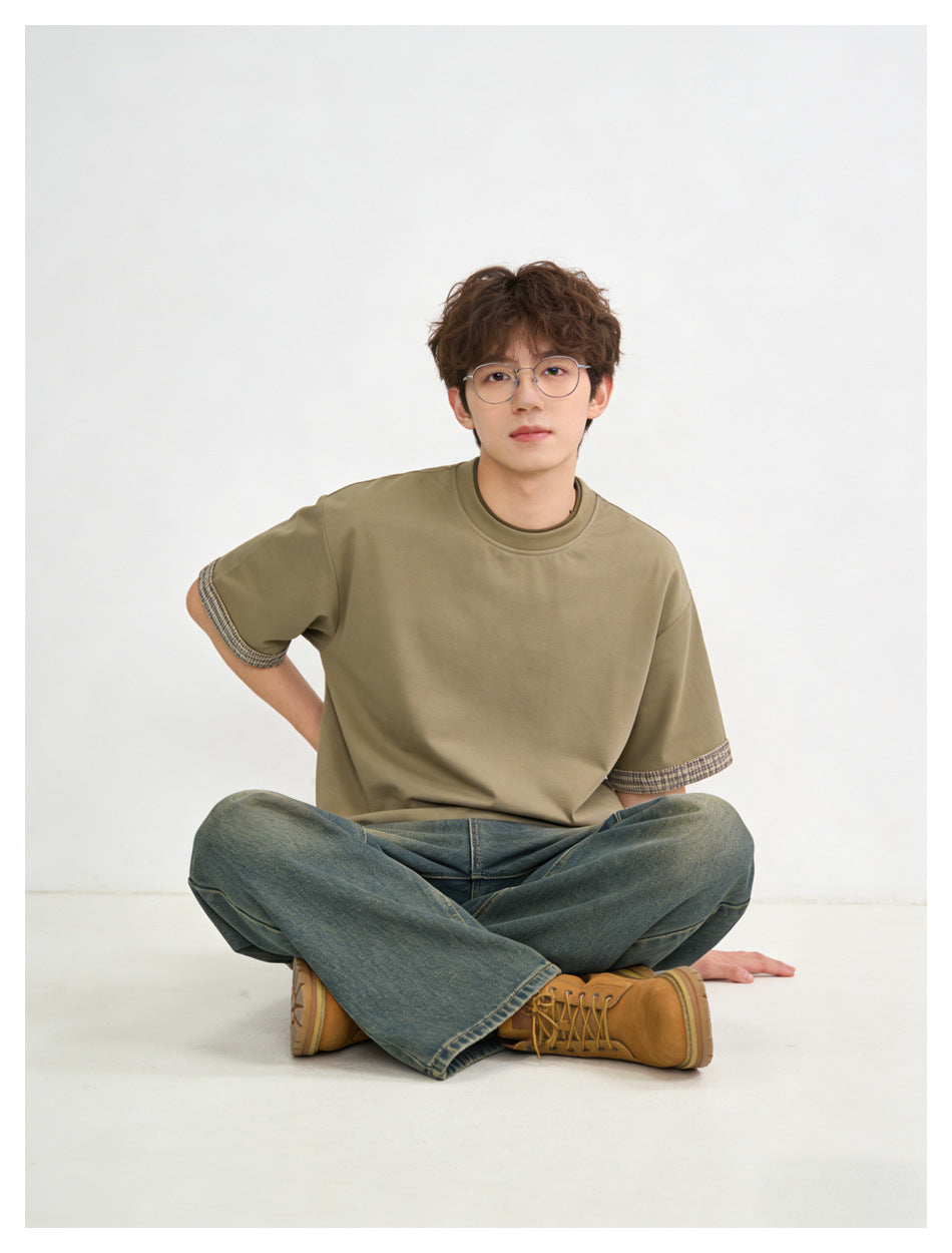 Seated model wearing an olive-brown short-sleeve T-shirt with patterned sleeve cuffs and loose blue jeans.
