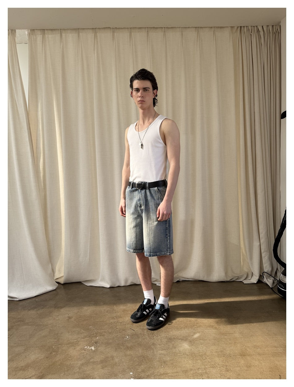 Model in a three-quarter pose wearing knee-length washed blue denim shorts with a white tank top, belt and sneakers