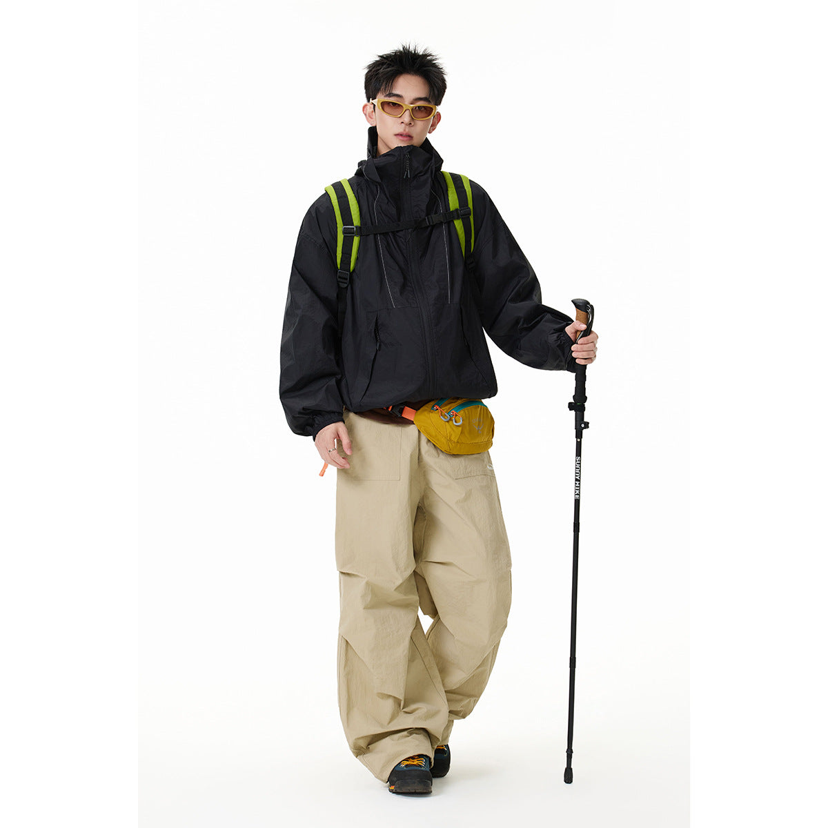 Model wearing a black high-collar jacket and tan trousers with a backpack, sunglasses, and trekking pole.