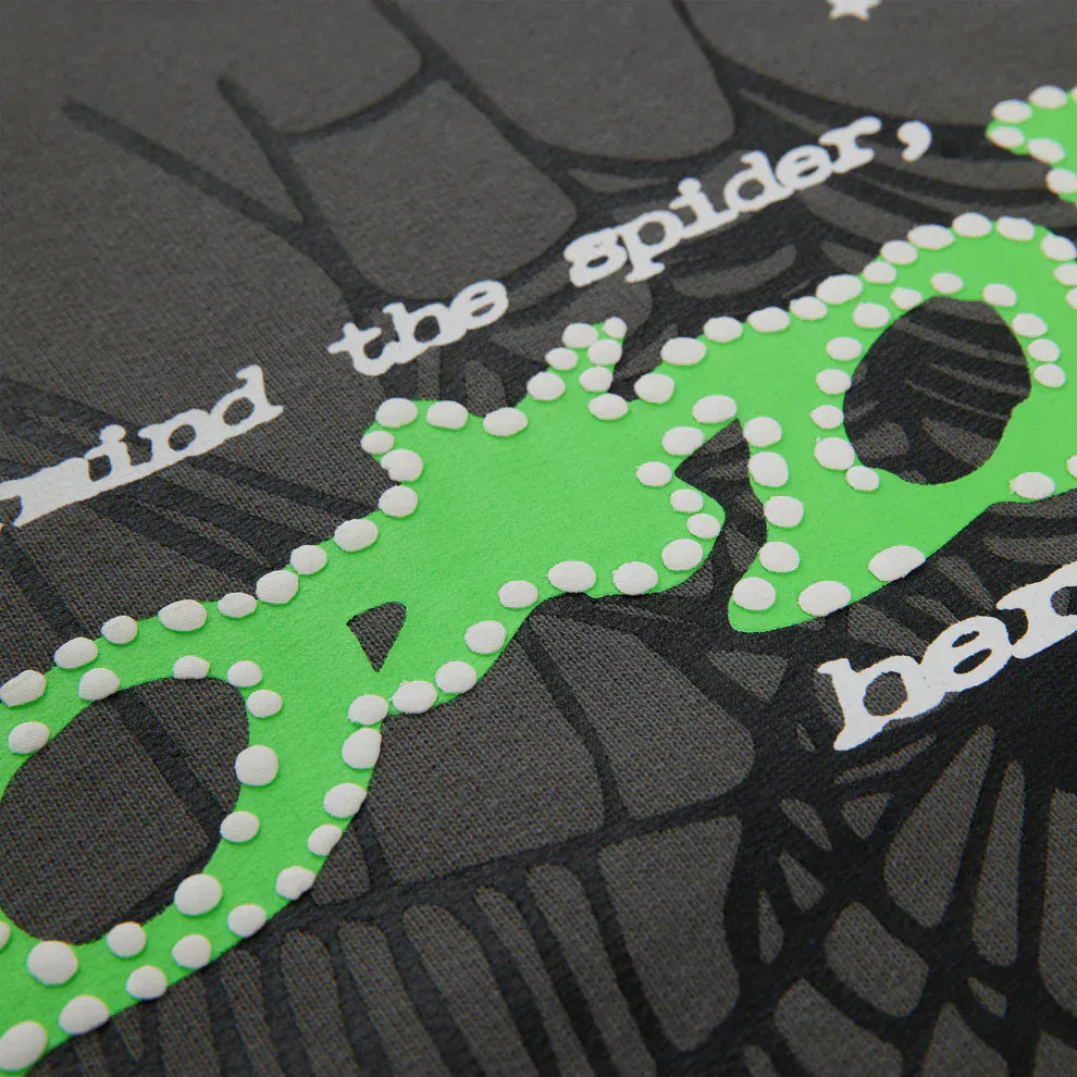 Close-up of graphic text and designs on the Sp5der “P!nk! Hares the Slime” Hoodie—an olive green 440GSM heavyweight pullover with white and green lettering, dotted outlines, and a dark spiderweb background.