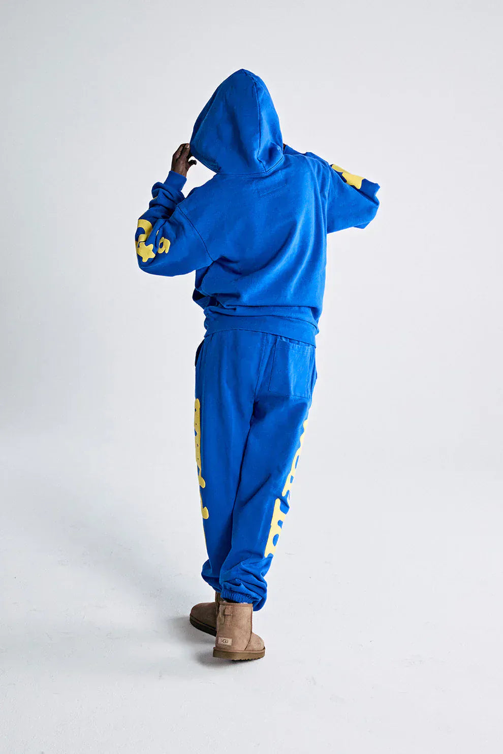 A person faces away from the camera in a Sp5der 440GSM Heavyweight Hoodie – Royal Blue with Yellow Star 3D Puff Print, matching sweatpants, and tan boots, channeling a Y2K streetwear vibe against a plain white background.