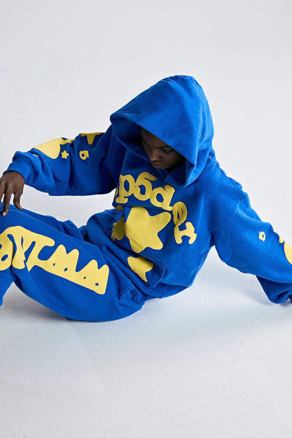 Person wearing a Sp5der 440GSM Heavyweight Hoodie in royal blue with yellow star 3D puff print sits on a white floor, looking downward in true Y2K streetwear style.